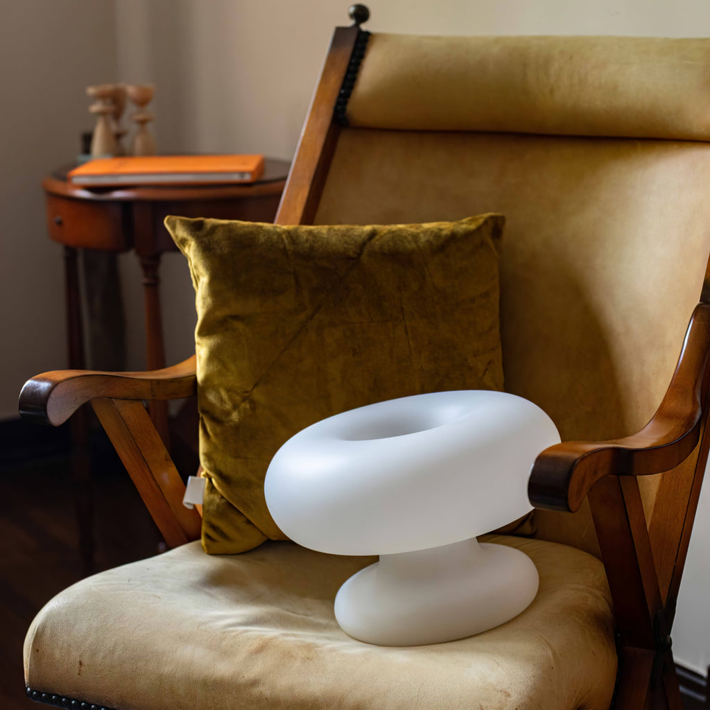 Clérachic donut lamp resting on the seat of a wooden armchair, with a mustard velvet cushion and a small round table behind.