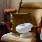 Clérachic donut lamp resting on the seat of a wooden armchair, with a mustard velvet cushion and a small round table behind.