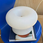 Clérachic donut lamp perched on a large book on a blue chair, its frosted shade wide above the narrower base.