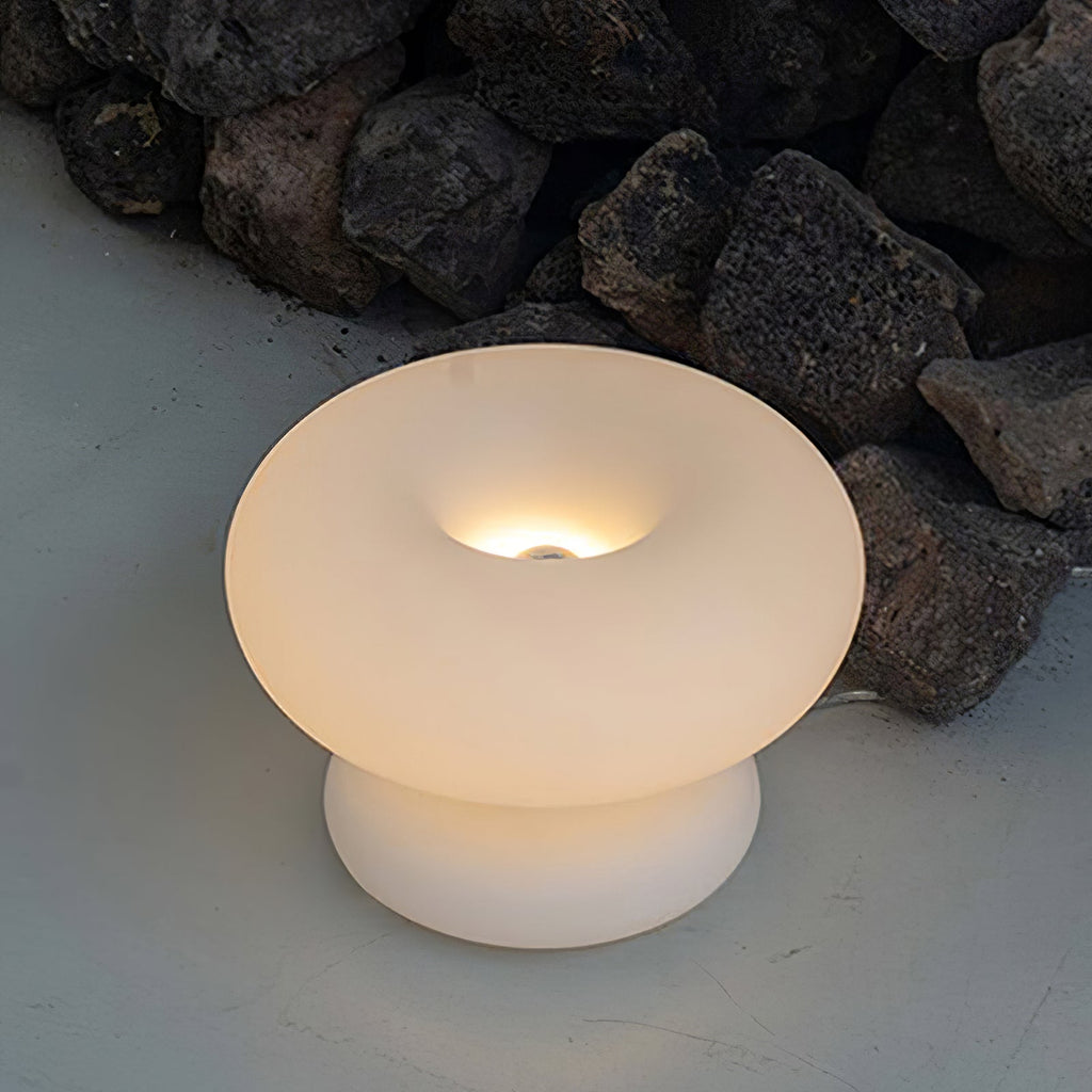 Lit Clérachic donut lamp on a pale surface in front of dark volcanic rocks, the bulb glowing inside the ring.
