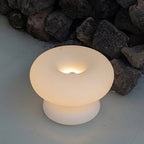 Lit Clérachic donut lamp on a pale surface in front of dark volcanic rocks, the bulb glowing inside the ring.