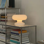 A Clérachic donut lamp glowing warmly on a chrome shelving unit, among stacked books and magazines.