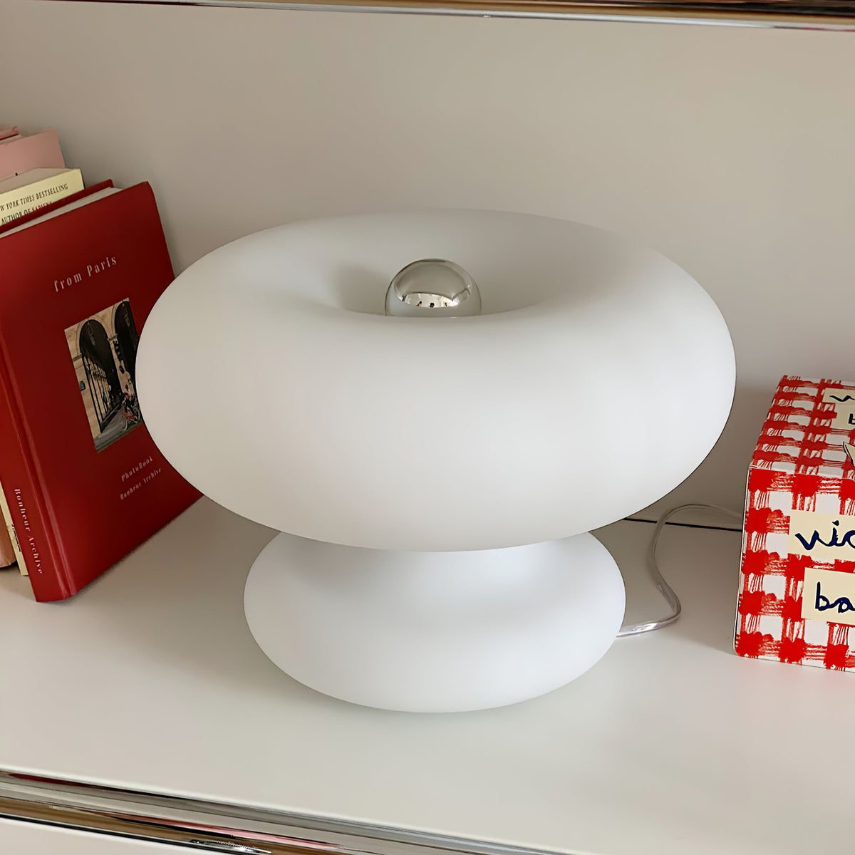 Close view of this Clérachic donut lamp on a white shelf, with red books and a checked box beside it.