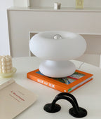 White Clérachic donut lamp on an orange book on a round white table, with a bobbled candle and a black arched ornament.