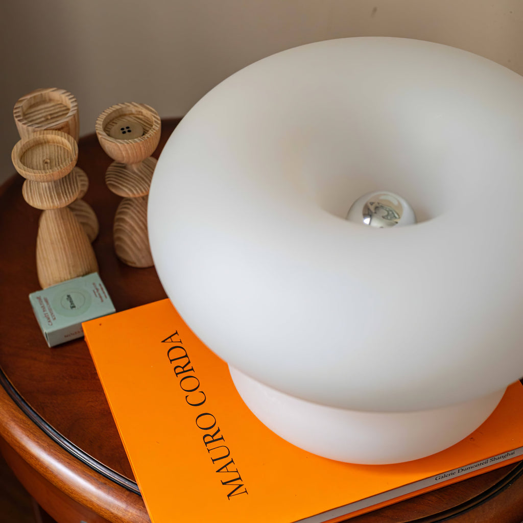 Close, angled view of the donut-shaped frosted shade of this Clérachic lamp, sitting on an orange book beside wooden candlesticks.