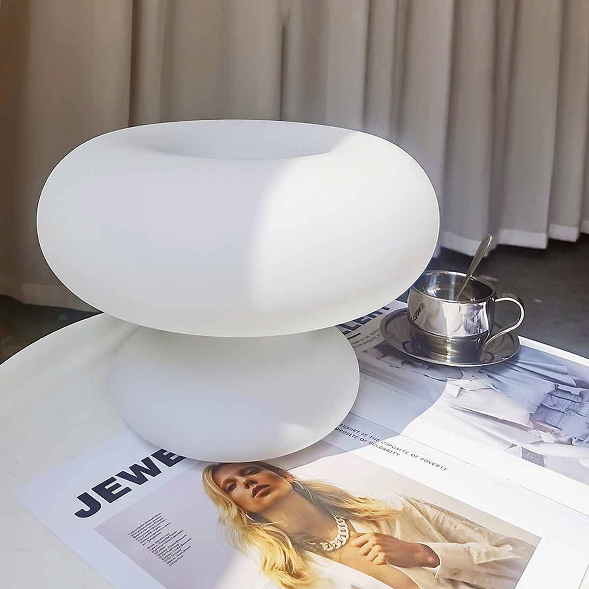 Clérachic donut lamp standing on a magazine on a glass table, with a silver tea cup and saucer and grey curtains behind.