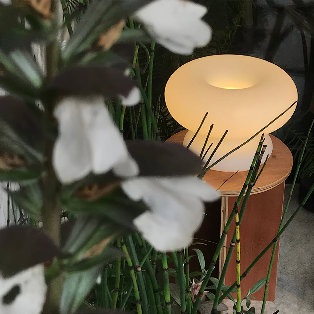 Lit Clérachic donut lamp glowing amber behind the dark leaves and white flowers of a plant, on a small wooden stool.