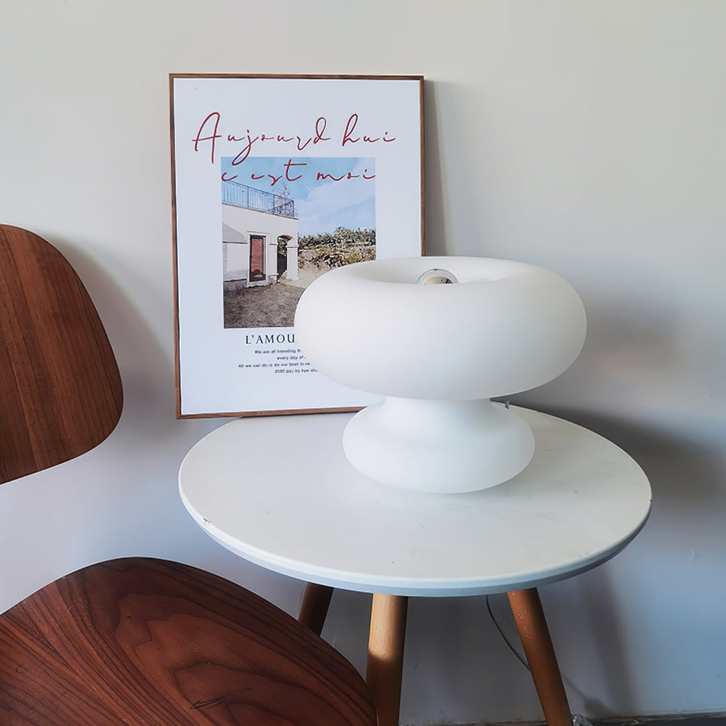 White Clérachic donut lamp on a round white table with wooden legs, in front of a framed poster and a walnut chair.