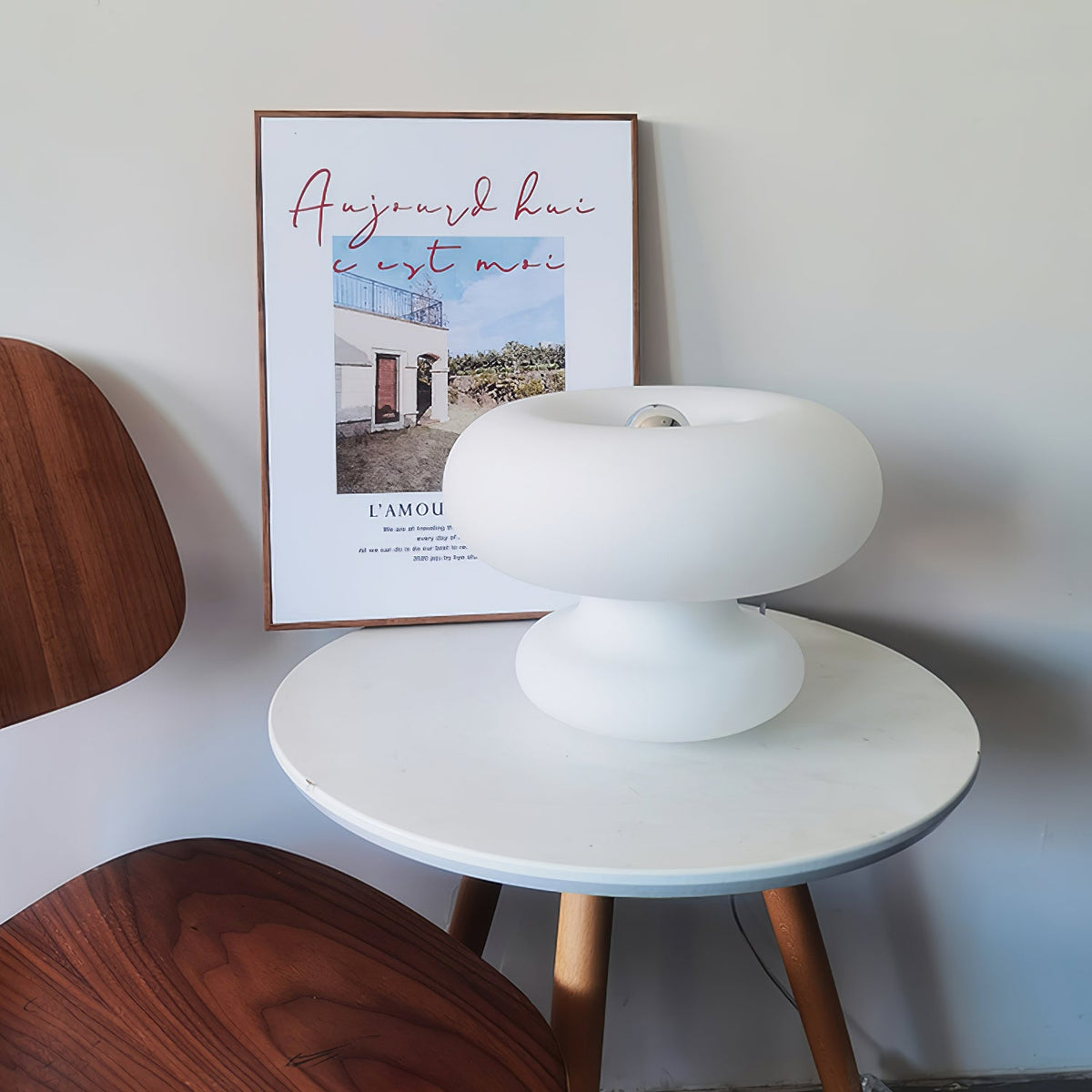 White Clérachic donut lamp on a round white table with wooden legs, in front of a framed poster and a walnut chair.