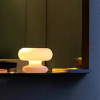 A Clérachic donut lamp glowing yellow on a dark shelf in front of olive-green and navy panels, with a book beside it.