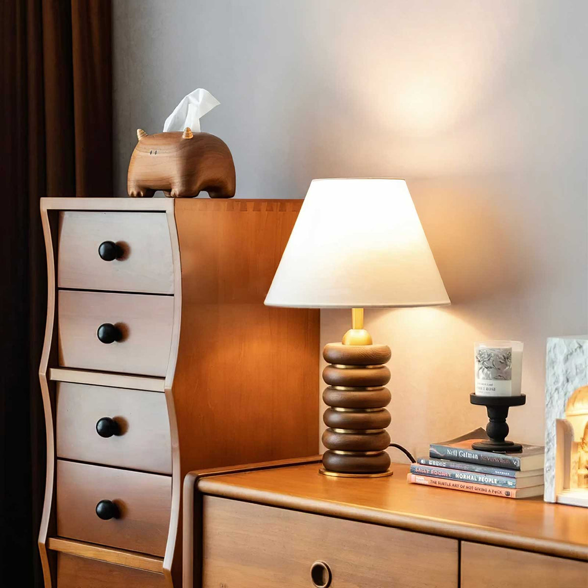 A Clérachic table lamp lit on a wooden chest of drawers, beside a curved drawer tower, books and a candle on a stand.