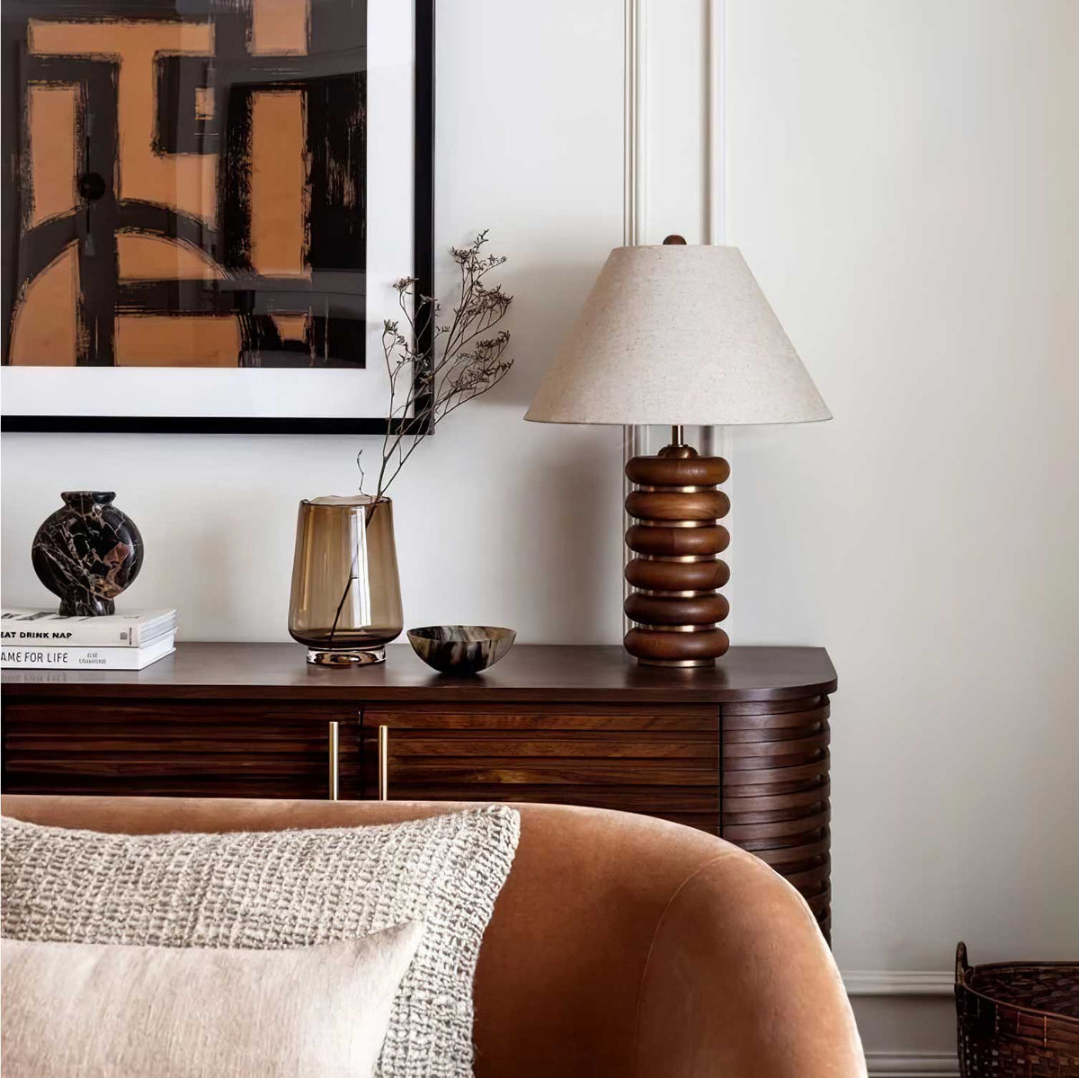 A Clérachic table lamp stands on a wooden sideboard with a glass vase and dark ornament, beside a large framed print.