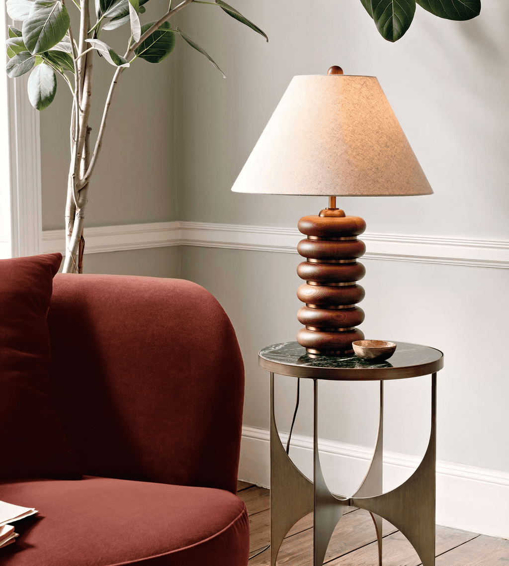 A Clérachic table lamp on a slim round table with metal legs, beside a rust-red armchair and a tall leafy plant.