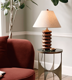 A Clérachic table lamp on a slim round table with metal legs, beside a rust-red armchair and a tall leafy plant.
