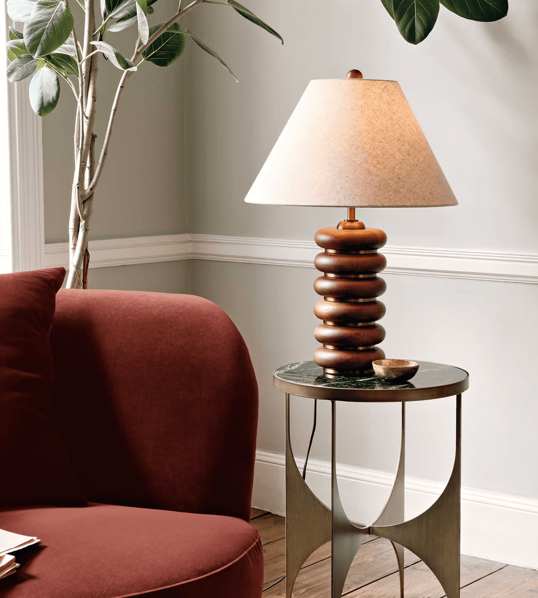 A Clérachic table lamp on a slim round table with metal legs, beside a rust-red armchair and a tall leafy plant.