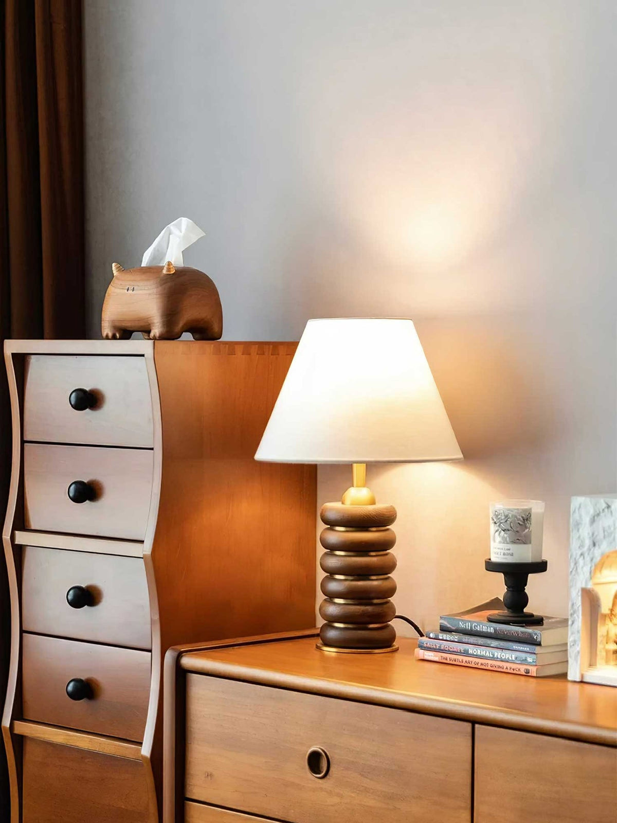 A Clérachic table lamp lit on a wooden chest of drawers, with a curved drawer tower, a candle and books beside it.