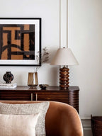 A Clérachic table lamp on a ribbed wooden sideboard, with a framed abstract print above and a tan velvet armchair in front.