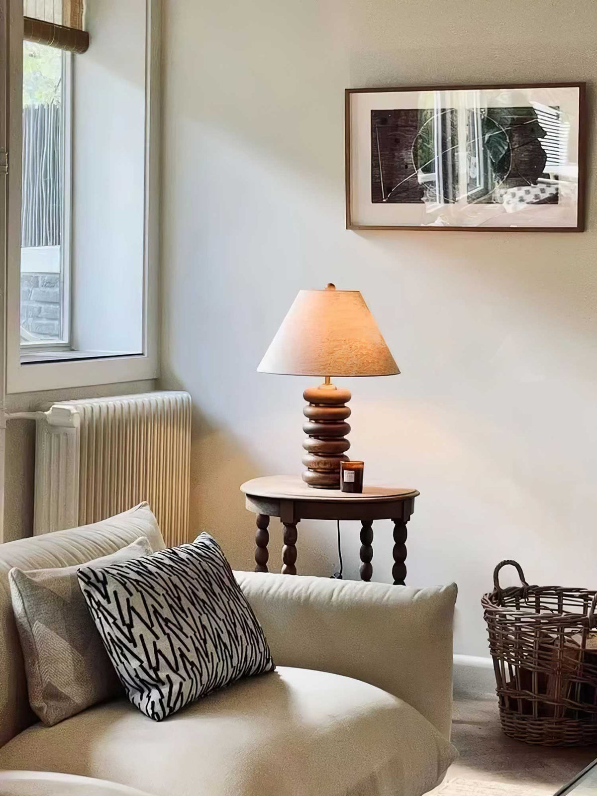 A Clérachic table lamp glows on a round turned-leg table in a corner with a cream sofa, a radiator and a framed print.