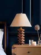 A Clérachic table lamp on a wooden bedside chest in a deep navy room, with a candle, a glass and dried flower stems.