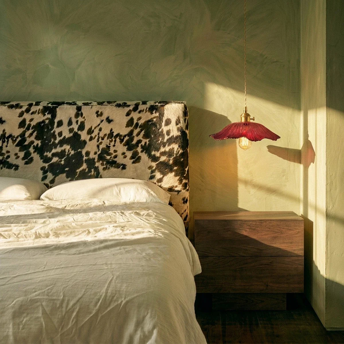 Clérachic red glass pendant with a flared shade hung beside a walnut bedside unit, in a bedroom with sage plaster walls and low sun.