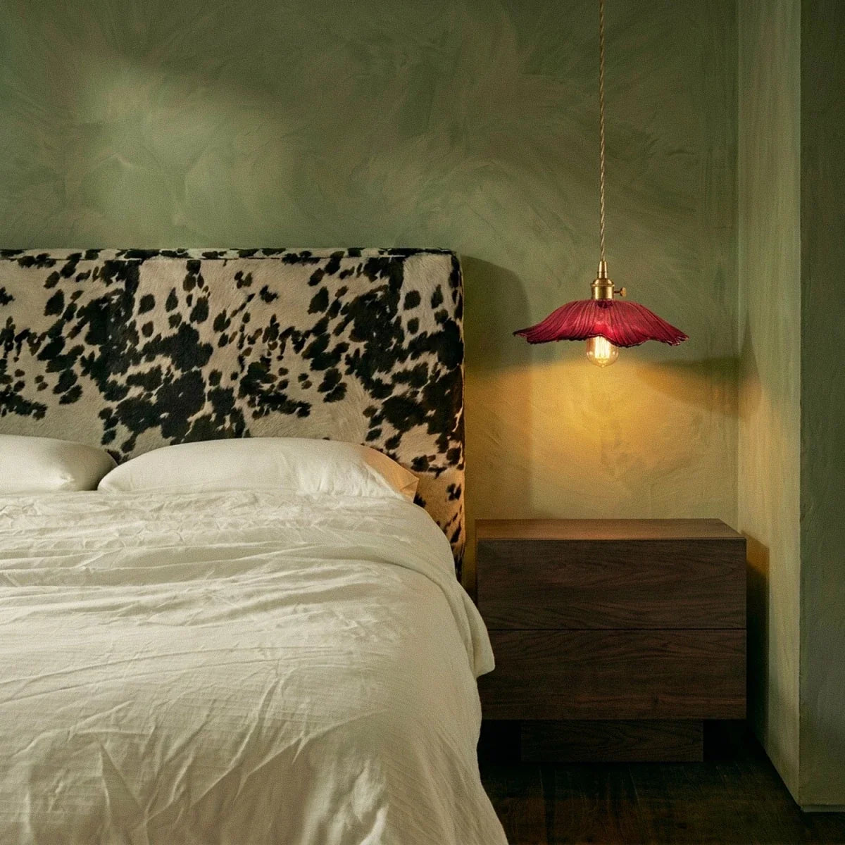 Clérachic red flared glass pendant lit at dusk beside a walnut bedside unit, with a cow-print headboard and sage plaster wall.
