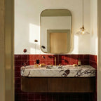 Clérachic clear ribbed glass pendant glowing above a marble basin, with a gilt mirror and burgundy tiles in a bathroom.
