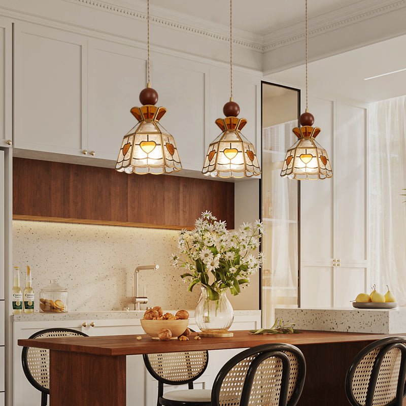 Three Clérachic stained glass pendants with wooden finials hanging over a kitchen island, with cream cabinets and a vase of white flowers.