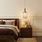 A Clérachic pendant with a frosted glass cage shade and a wooden finial hung beside a dark wood bed and a bedside table with books.