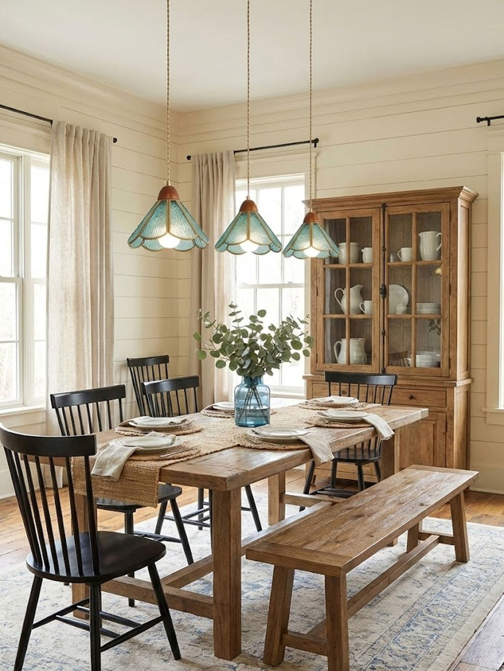 Three teal Clérachic stained glass pendants above a farmhouse dining table, with black chairs, a bench and a glazed wooden hutch.