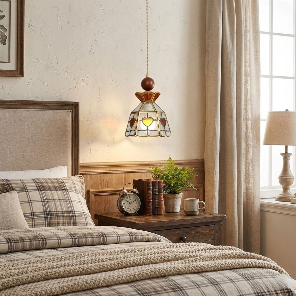 A Clérachic stained glass pendant with amber hearts hanging beside a bed with plaid bedding, a stack of books and a small plant.