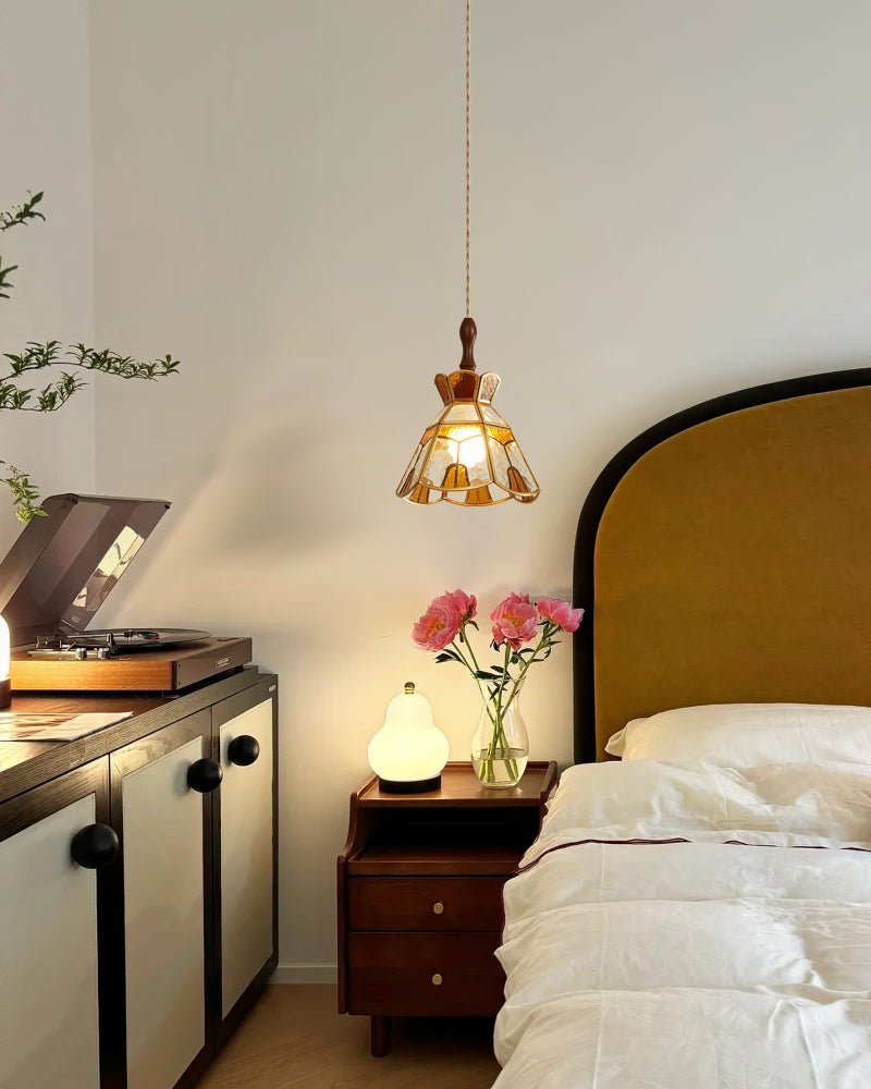 A Clérachic heart-panel pendant lit beside a bed with a mustard headboard, a bedside table with pink flowers and a record player.