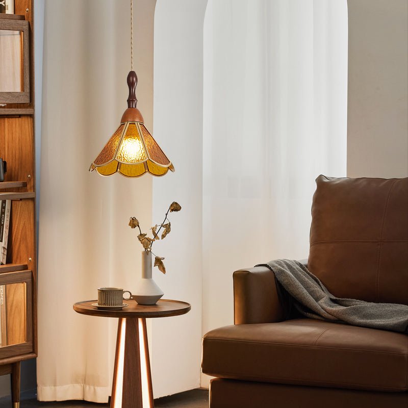 An amber Clérachic pendant with a scalloped shade by a sheer curtain, with a leather armchair, a grey throw and a round side table.