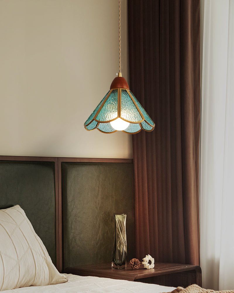 A teal Clérachic pendant glowing above a bed with a dark green panelled headboard and a heavy brown curtain beside it.