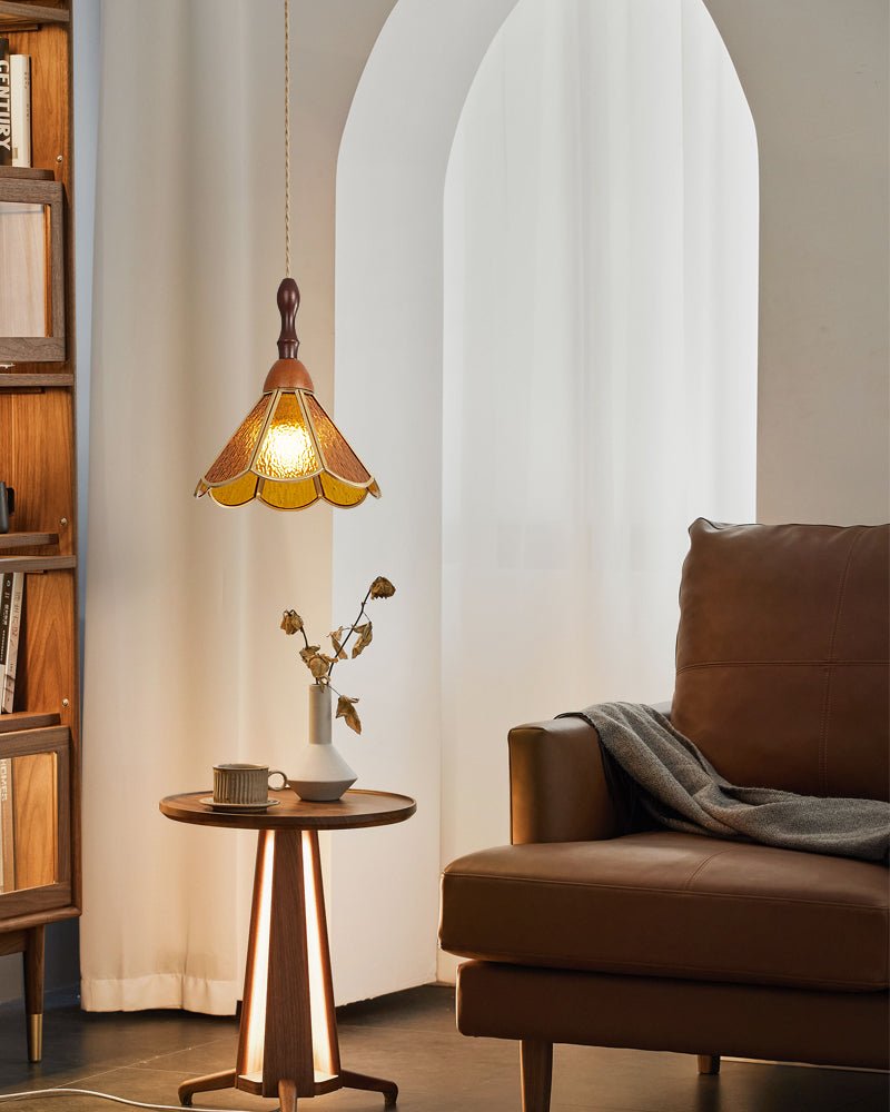 An amber Clérachic pendant with a scalloped glass shade, lit beside a tall arched curtain, a brown armchair and a small table.