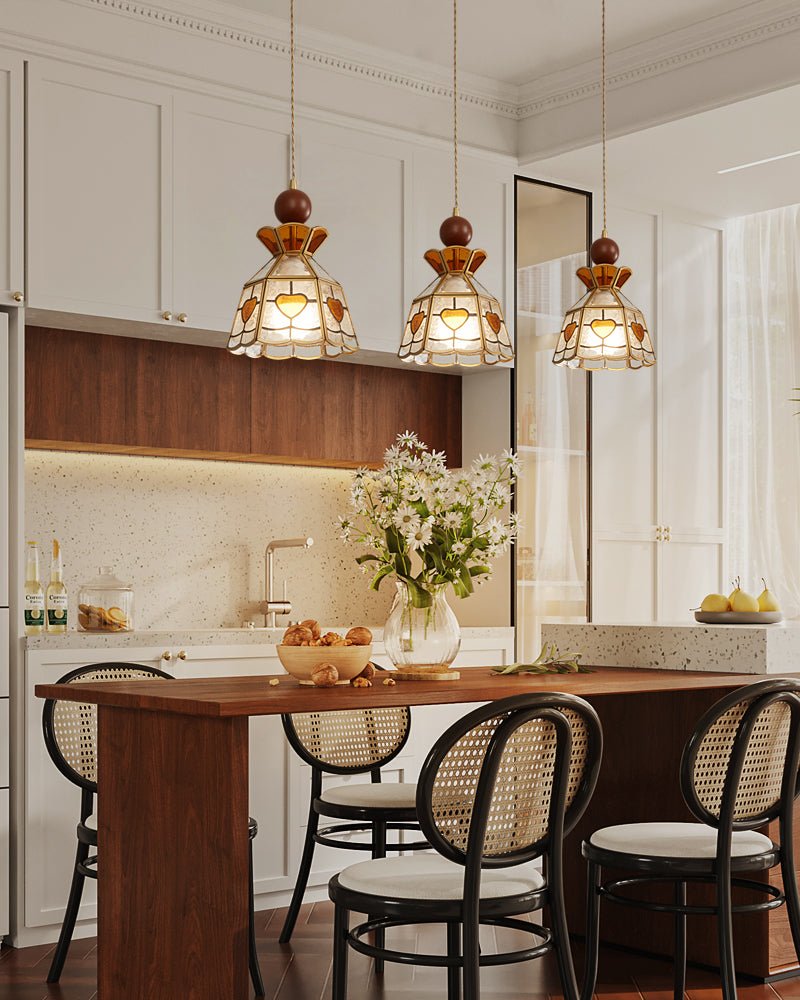 Three Clérachic heart-panel pendants over a dark wooden dining table, with cane-backed chairs and a vase of flowers beneath them.