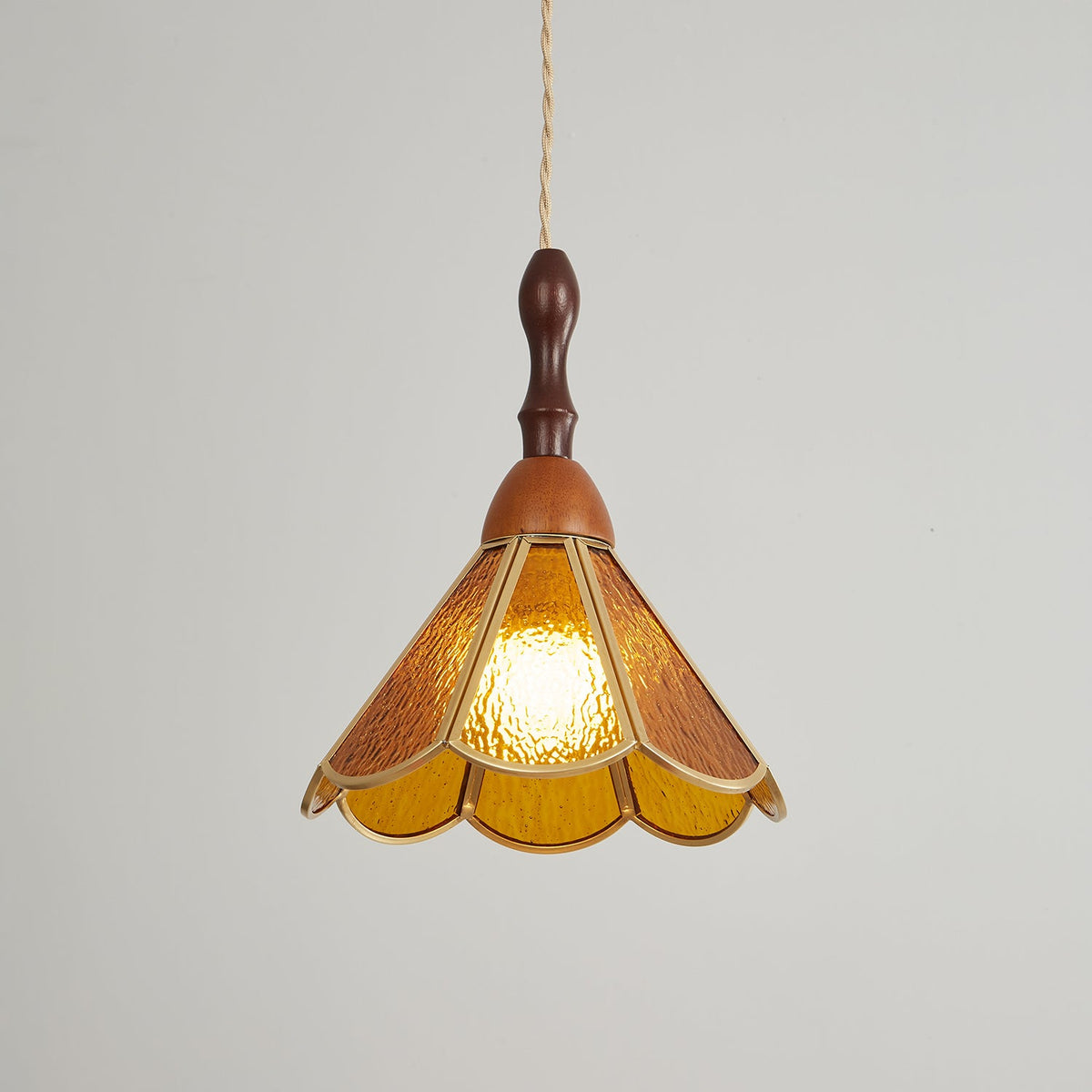 Amber Clérachic pendant on a grey backdrop, its textured glass petals edged in brass and lit from within by a warm bulb.