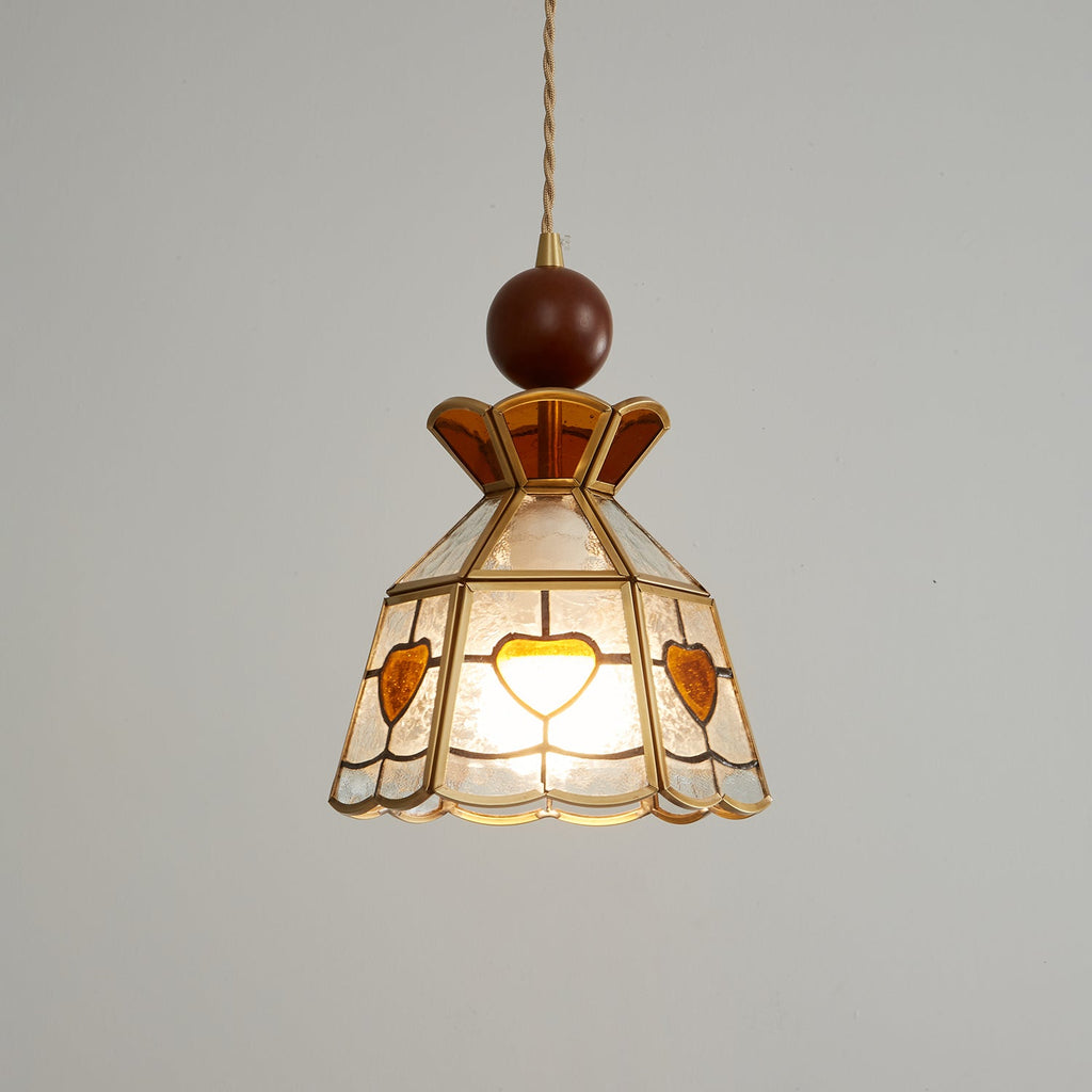 Closer view of the lit Clérachic heart-panel pendant, its amber crown and brass ribs sharp against a pale grey wall.