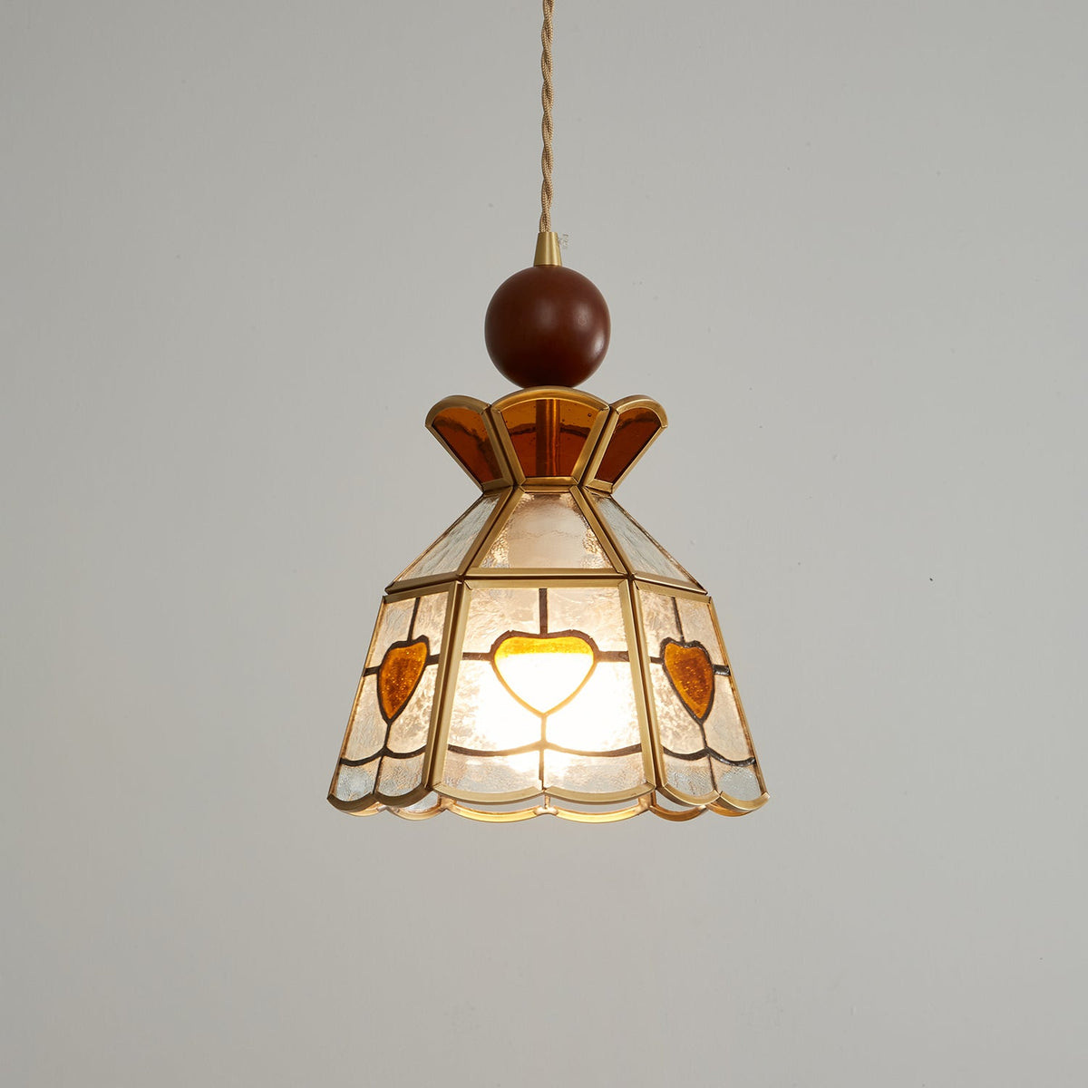 Closer view of the lit Clérachic heart-panel pendant, its amber crown and brass ribs sharp against a pale grey wall.