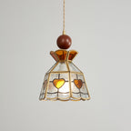 Clérachic heart-panel pendant seen at a slight angle, lit warm, with amber hearts and a wooden ball finial on grey.