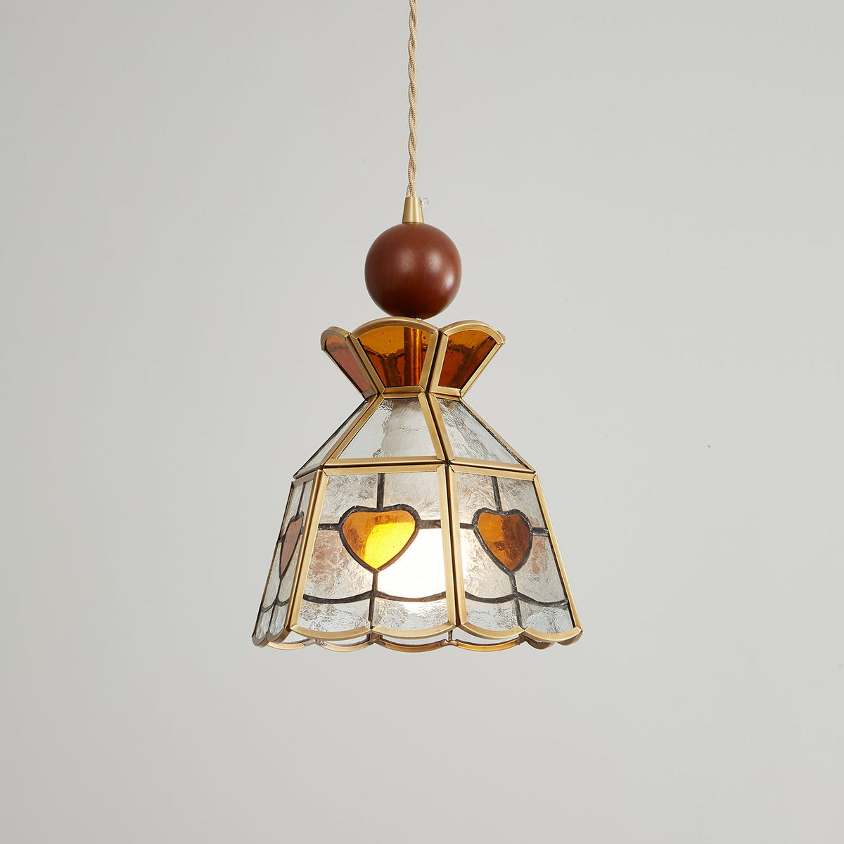 Clérachic heart-panel pendant seen at a slight angle, lit warm, with amber hearts and a wooden ball finial on grey.