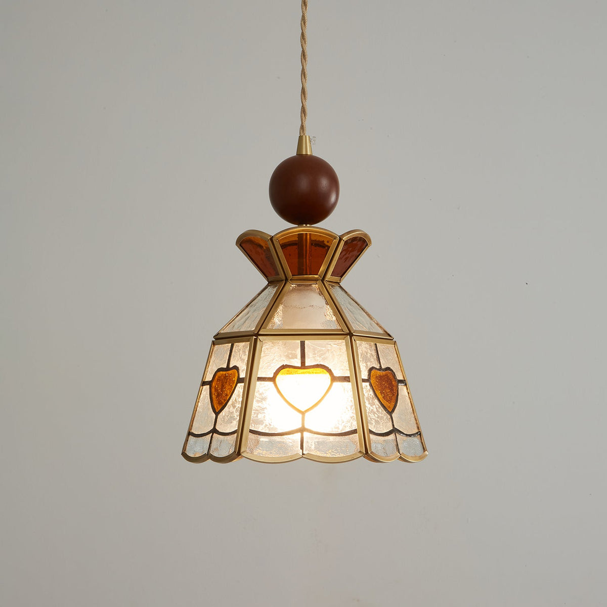 Clérachic heart-panel pendant, lit, with its wooden ball and brass fitting at the top, photographed on a grey backdrop.