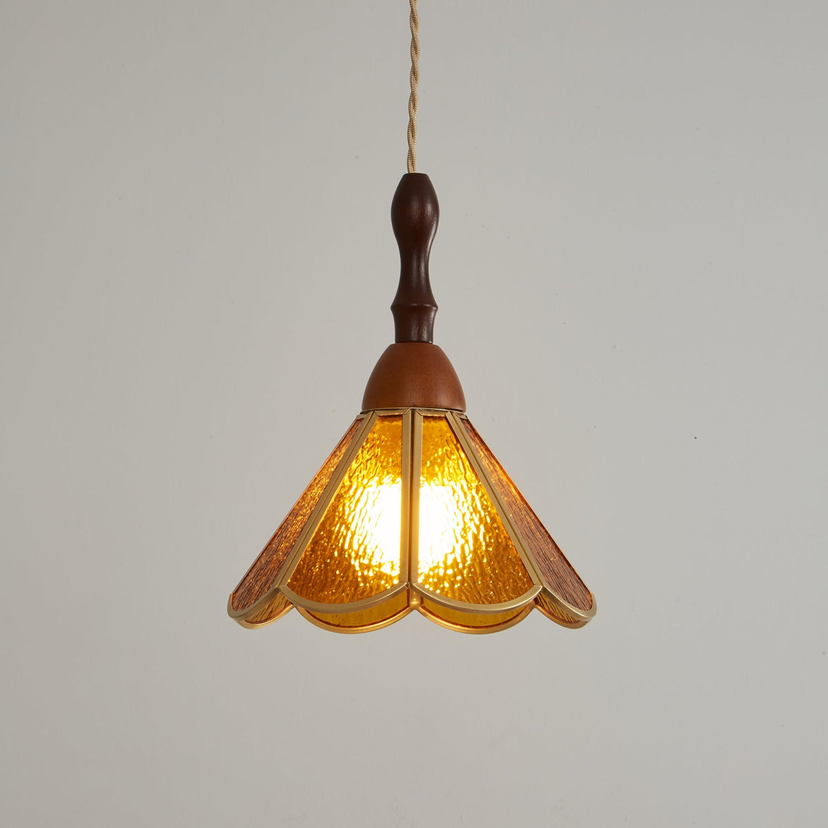Amber Clérachic pendant lit on a grey backdrop, a warm glow through the petal-shaped glass panels and a wooden cap above.