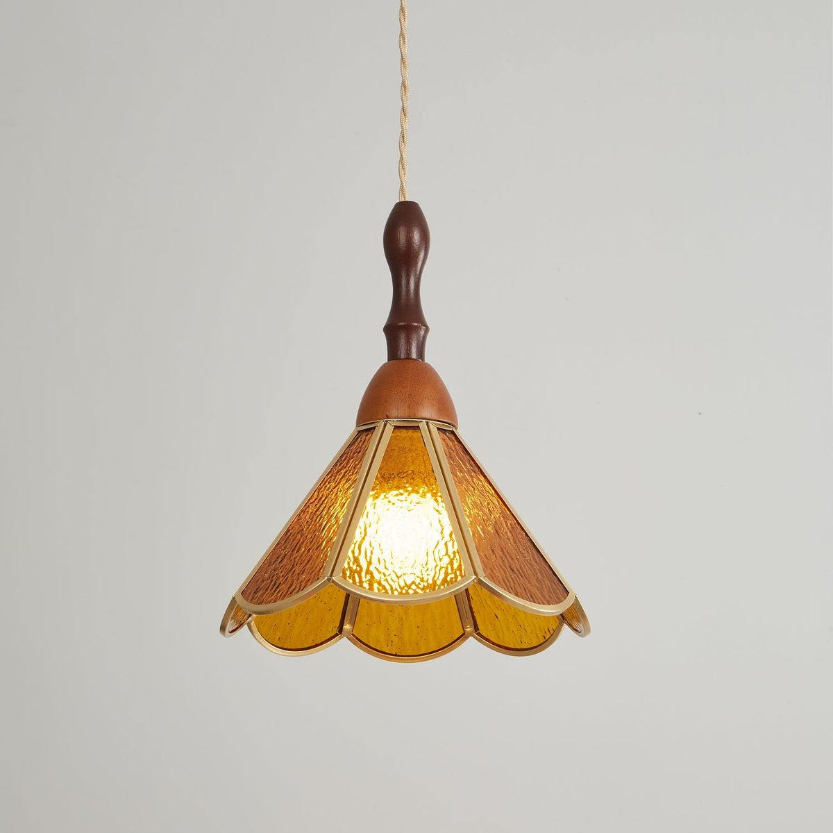 Amber Clérachic pendant lit on a pale backdrop, its scalloped shade glowing evenly and its wooden finial dark above.