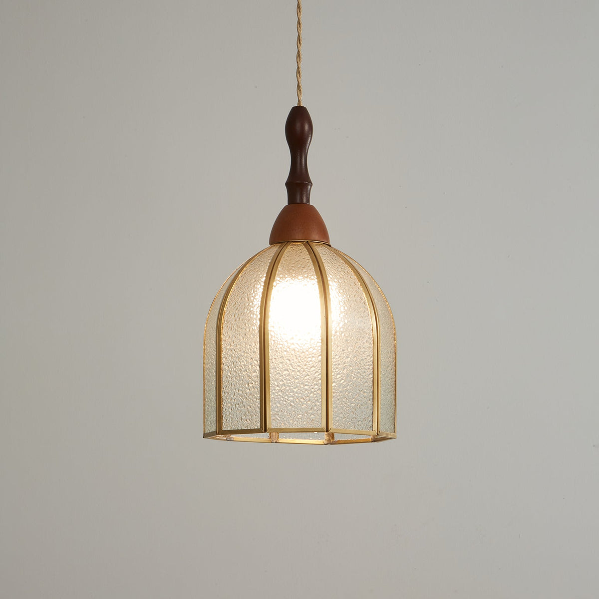 Lit Clérachic domed pendant glowing a soft gold, its brass ribs and textured glass clear against a grey wall.
