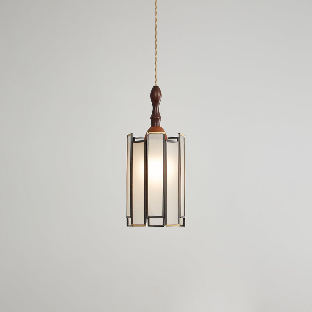 Lit Clérachic pendant with a tall frosted shade, the bulb glowing evenly behind panels in a dark metal frame.