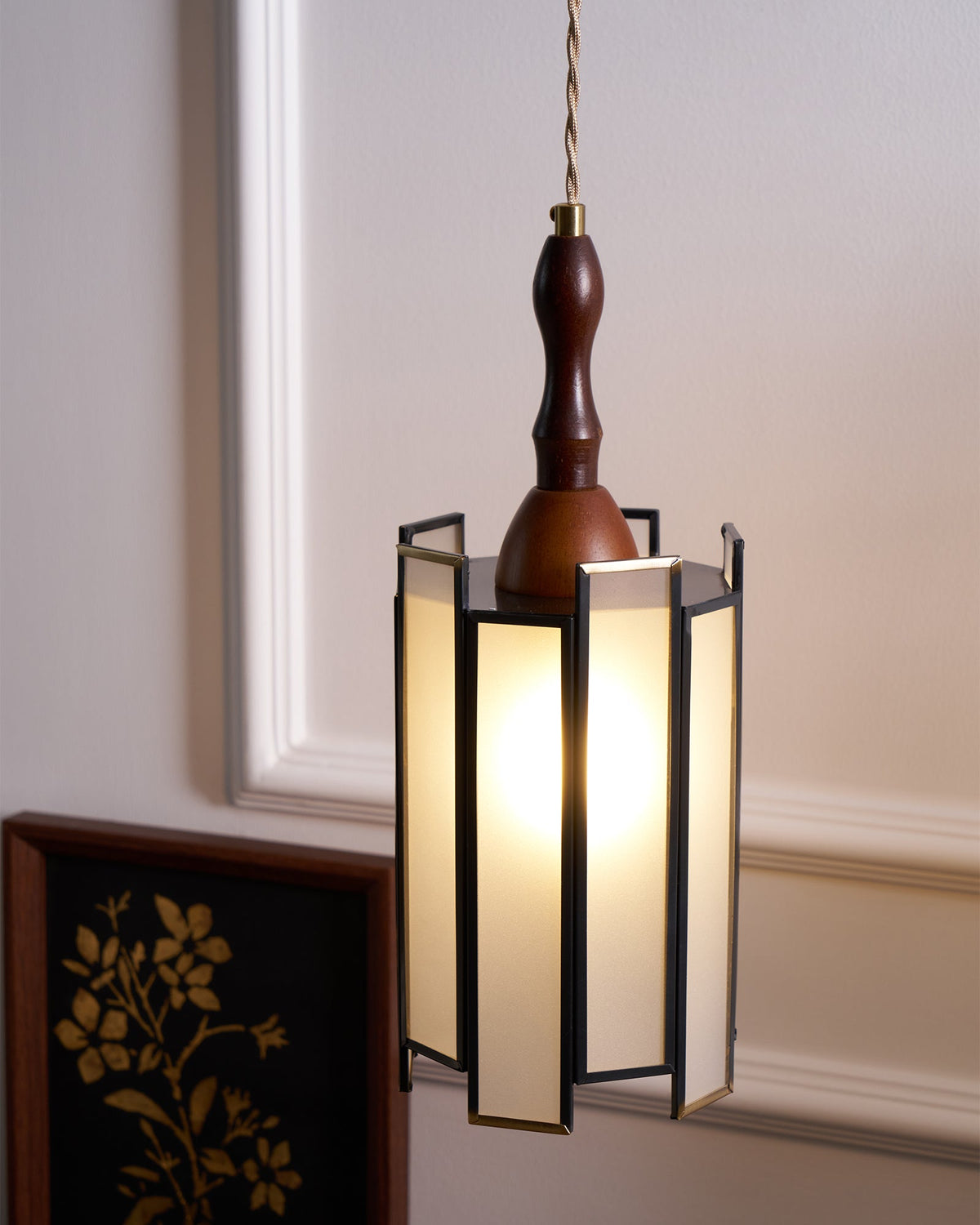 Lit Clérachic frosted-panel pendant beside a framed gold botanical print, against a cream panelled wall.