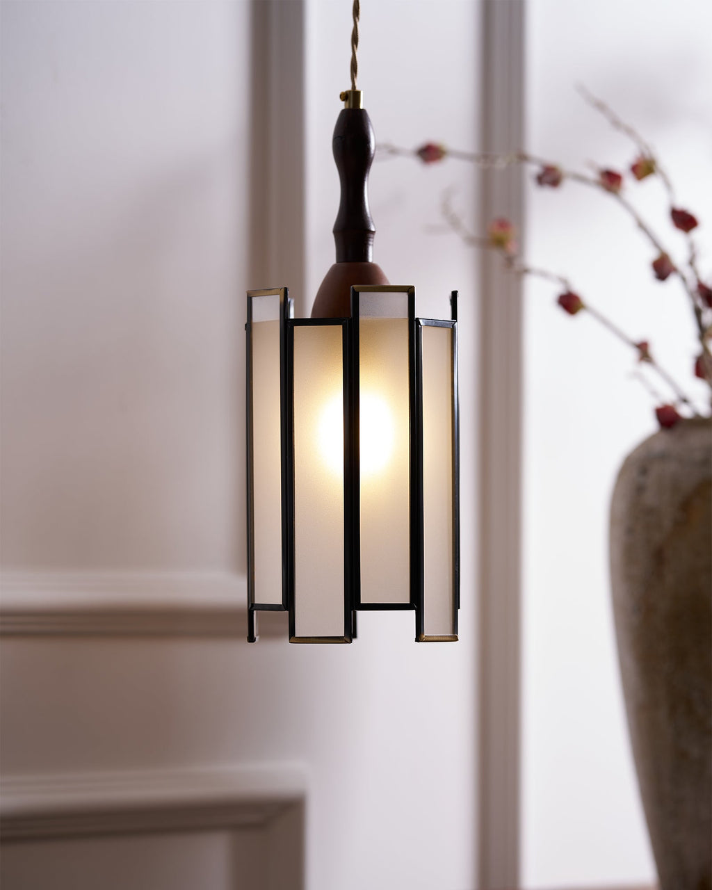 Clérachic frosted-panel pendant glowing in front of a stoneware vase with dried blossom branches, with wall panelling behind.