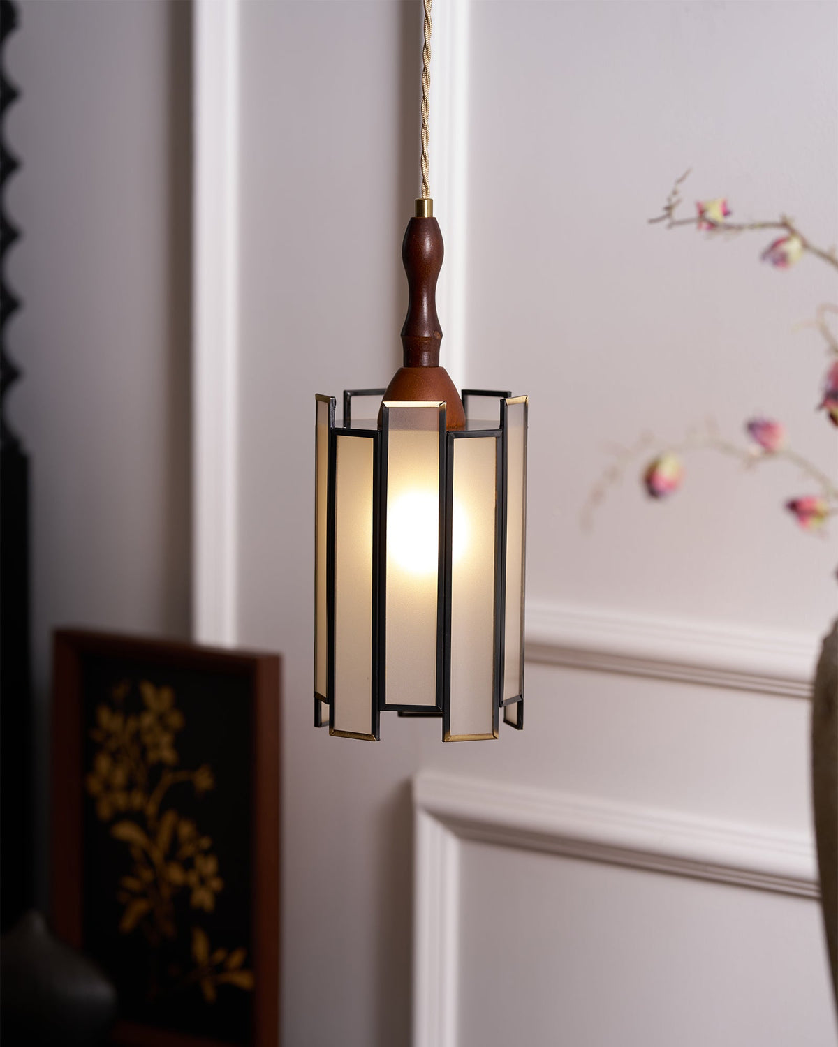 Clérachic frosted-panel pendant lit beside a framed gold botanical picture and a vase with blossom, in soft side light.