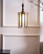 A small Clérachic frosted-panel pendant glowing on a cream panelled wall, with a blossom branch above and a dark table below.