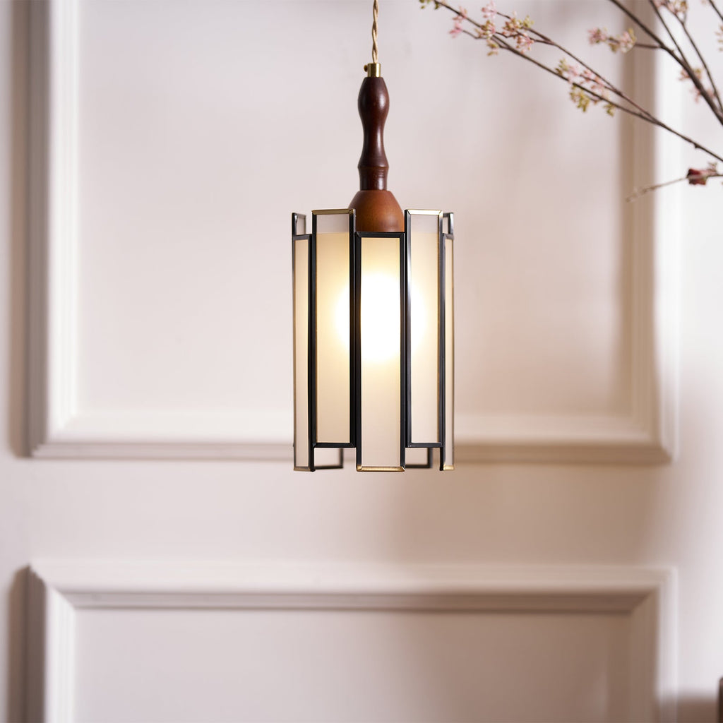 A lit Clérachic frosted-panel pendant hung against a panelled wall, with a blossom branch reaching in from the top.